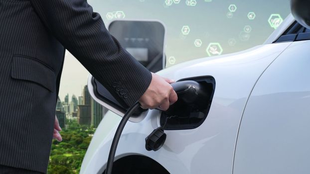 Progressive green city ESG symbol background with electric vehicle. Businessman recharge EV car's battery with charger plug at charging station in the urban with ecological and green symbols signs.