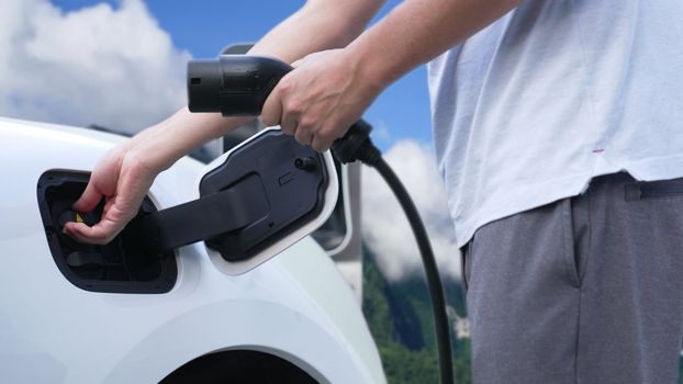 Progressive man recharge electric vehicle installing power cable plug from charging station with rural mountain in the background. EV car driven by electro engine for clean environment3