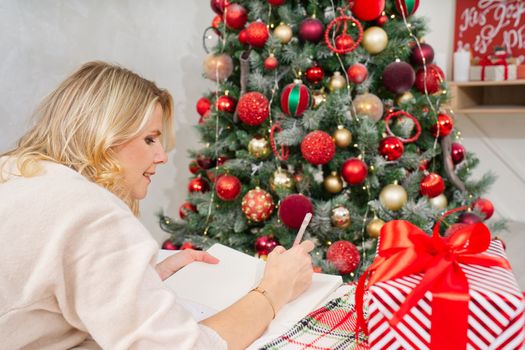 happy woman with pen and notebook making wish list or to do list for new year in bed over christmas tree. woman dreaming in christmas time. xmas holidays concept