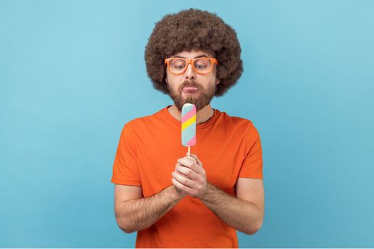 Portrait of man with Afro hairstyle wearing orange T-shirt and funny eyeglasses, holding ice cream, feels hungry, wants to lick dessert. Indoor studio shot isolated on blue background.