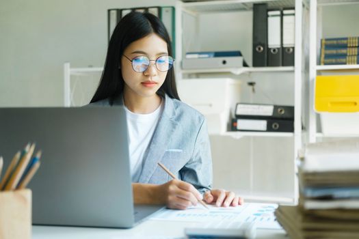 Young asian businesswoman working hard at the office using laptop data graphs, planning for improvement, analyzing and strategizing for business growth. Business concept