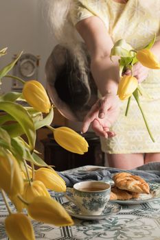 Drinking coffee and tea, a middle-aged blonde woman with long hair drinks tea from a vintage cup and eats puff pastry with pecans on the background of yellow tulips and books on the table