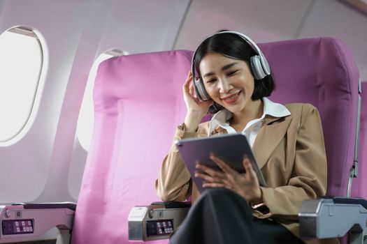 Young caucasian smiling female enjoying her comfortable flight while sitting in airplane cabin, listening to music in earphones and drinking water. Wifi internet access on board, passenger near window..