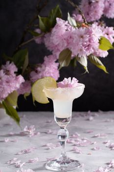Spring still life with a glass of cold Margarita with lime surrounded by pink sakura flowers on a cement backgroundrefreshing summer drink alcoholic non-alcoholic cocktail. High quality photo