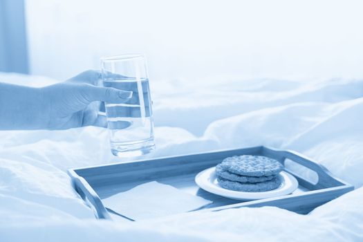 Female hand holding glass of water over tray with crackers on a bed