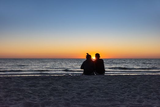 St. Petersburg, Florida, USA - Oct. 22, 2022: Beach lovers watching the sunset sitting on a sandy beach.  Beach scene with couple silhouette. Romantic couple on the beach. St. Petersburg, Florida