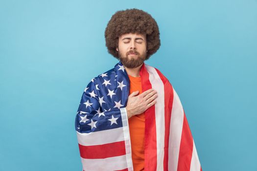 Serious man with Afro hairstyle wrapped USA flag, keeping hand on chest, keeps eyes closed, proud of his social security in country. Indoor studio shot isolated on blue background.
