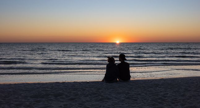 St. Petersburg, Florida, USA - Oct. 22, 2022: Beach lovers watching the sunset sitting on a sandy beach.  Beach scene with couple silhouette. Romantic couple on the beach. St. Petersburg, Florida