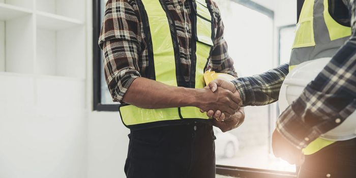 Engineers or architecture shaking hands at construction site for architectural project, holding safety helmet on their hands
