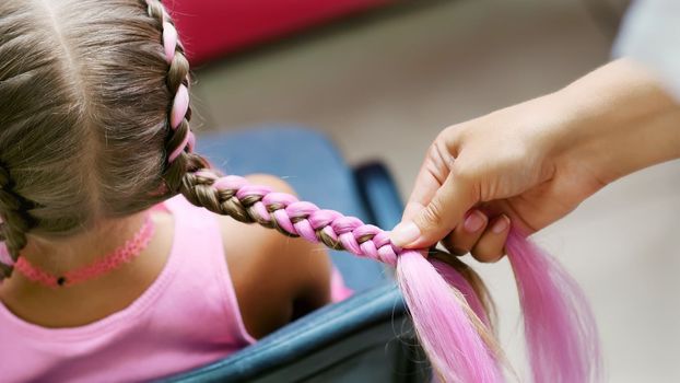 Beautiful blond girl, of seven years old, braided two pigtails, do a hairstyle with pink locks of hair in a beauty salon, a hairdresser's salon, in front of a large mirror. a little princess. High quality photo