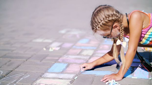 a girl in sunglasses, draws drawings with colored crayons on the asphalt, street tiles. A hot summer day. High quality photo