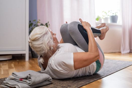 Senior woman lying on yoga mat doing apanasana. Fit female relaxing on floor at home. 