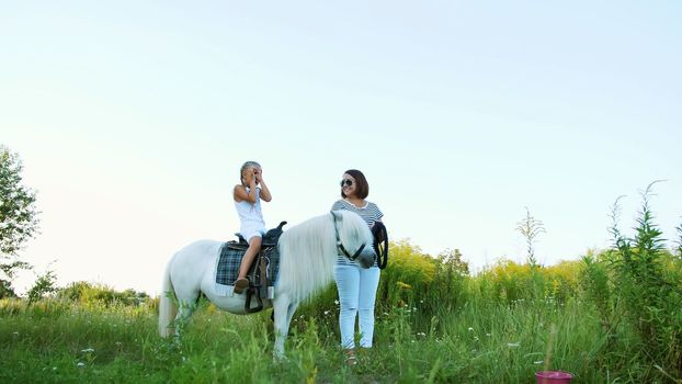 Mom and daughter are walking around the field, daughter is riding a pony, mother is holding a pony for a bridle. Cheerful, happy family vacation. Outdoors, in summer, near the forest. High quality photo