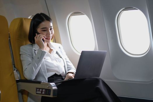 Asian woman passenger sitting in airplane near window and reading news from social networks or using travel applications in smartphone.