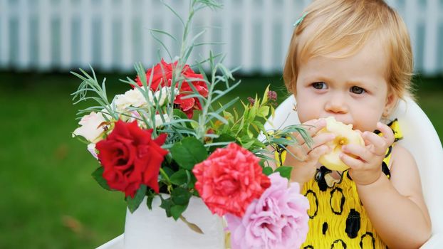 summer, in the garden. portrait, one year old pretty blond girl eating an apple with apetite. on the table, there is a beautiful bouquet of flowers. High quality photo