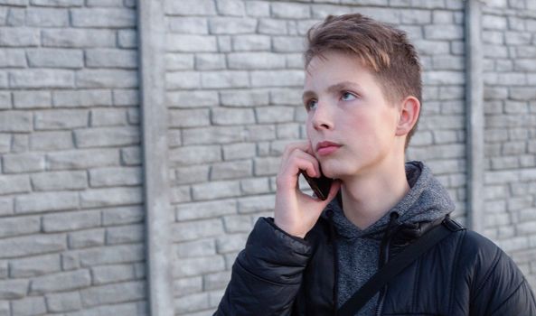 A teenage boy in a black jacket holds a smartphone in his hand against a background of a gray wall.
