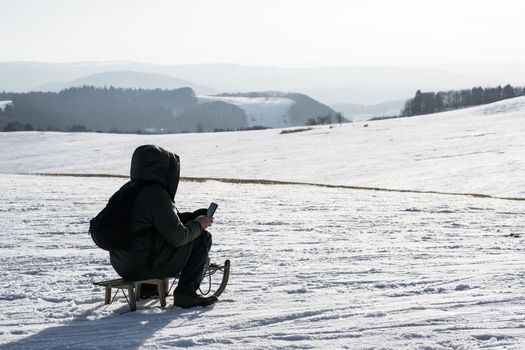 a man looks at his mobile phone sitting in the cold on top of a snowy mountain in winter on a children's sled wrapped in his winter warm jacket. High quality photo