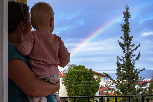 Mother looks at the sky with a baby in her arms, admires the rainbow after the rain, summer is outdoors. 