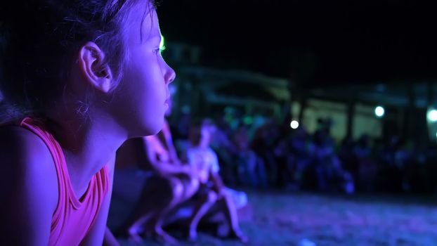 summer, night, in the rays of soffits, a girl of seven years old, watching the night concert in an open-air theater, on vacation. many children are in the background. High quality photo