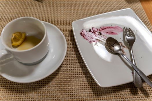 a dirty plate and an empty cup. Empty dishes after eating on a wooden table in a cafe. a white plate with traces of food and a cup with drunk tea and a piece of lemon. after breakfast. morning tea, snack