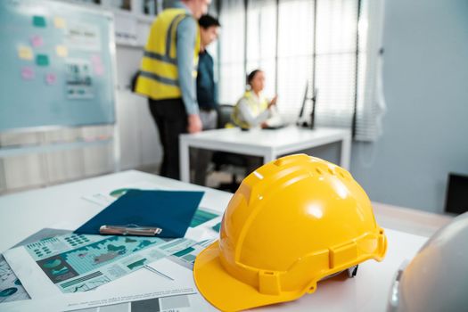 Concept of engineering equipments of protective helmet for competent engineer on piles of paper works. Blurred image of engineers and employer working together at the desk.