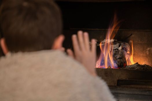 Rear view of young boy wrapped in a blanket warming hands in front of firewood in winter.