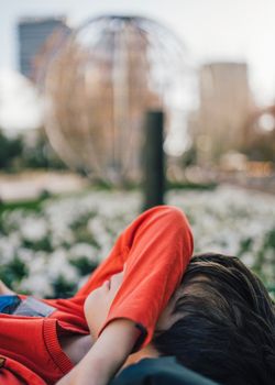 Side view of young boy lying in a city street park ground hiding face under arm covering eyes from light-