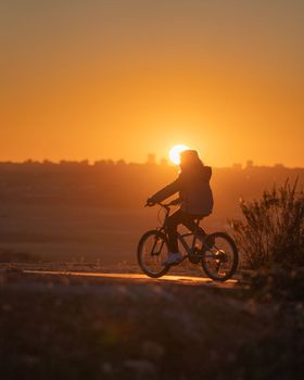 Silhouette of woman riding a bicycle against the sun and Madrid cityscape at sunset.