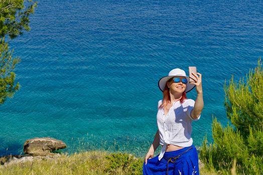 Smiling young woman in sunglasses and straw hat taking selfie against sea and blue sky.