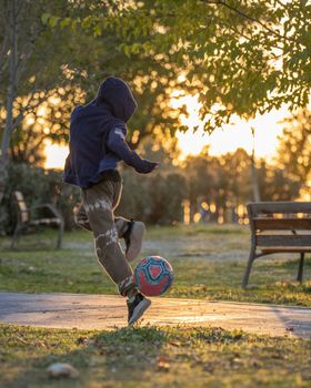 Young caucasian boy practicing soccer skills and tricks with the football ball at sunset in nature. Urban city lifestyle outdoors concept.