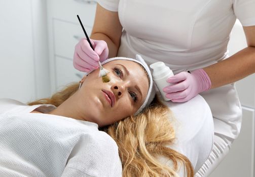 Cosmetologist applying a clay mask to the face of beautiful woman. Spa treatment in the beauty salon