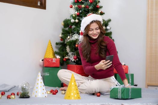 asian young woman has video chat on smartphone and shows present box to her friend with christmas tree.