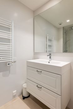 Spacious bright bathroom has washbasin with tap built into nightstand, large mirror that reflects shower cabin on opposite side of room and heating radiator for drying towels