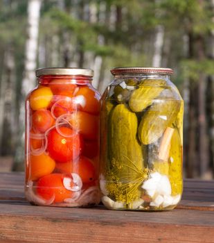 Two jars with pickled cucumbers and tomatoes are on a wooden table in nature.