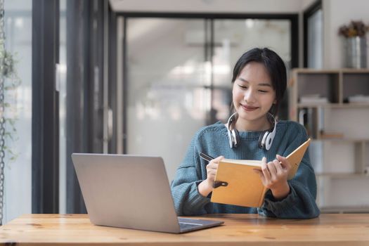 Young adult happy smiling Asian student wearing headphones talking on online chat meeting using laptop in university campus or at virtual office. College female student learning remotely