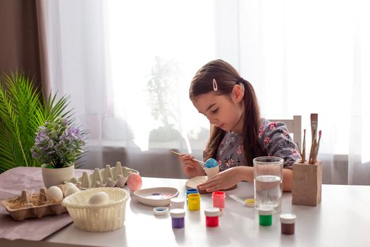 A cute little girl sits at a white table by the window, paints eggs with a brush and paint in jars. Copy space