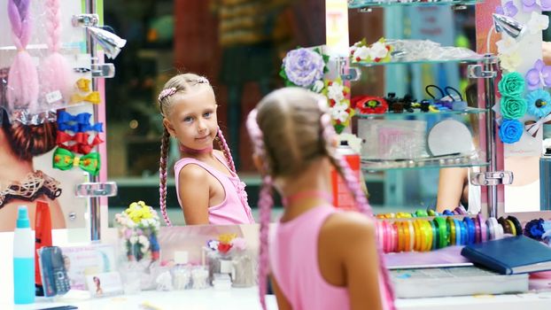 Beautiful blond girl, of seven years old, braided two pigtails, do a hairstyle with pink locks of hair in a beauty salon, a hairdresser's salon, in front of a large mirror. a little princess. High quality photo