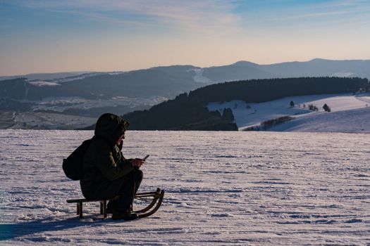 a man looks at his mobile phone sitting in the cold on top of a snowy mountain in winter on a children's sled wrapped in his winter warm jacket. High quality photo