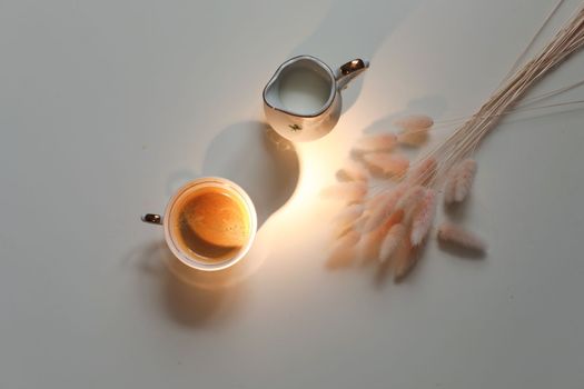 White cup with coffee and milk jug on a white wooden background, closeup. Energy breakfast, morning routine concept. coffee cup and milk pitcher on white table background