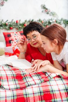 happy mother with daughter with pen and notebook making wish list or to do list for new year in bed over christmas tree. mother with daughter dreaming in christmas time. xmas holidays concept