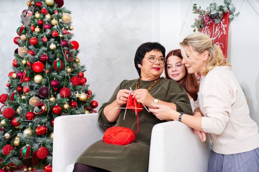 A cheerful 60 year old woman knitting a sock near the Christmas tree. elderly woman resting, relaxing during christmas holidays. grandmother, mother, daughter, son wife and granddaughter spending christmas time