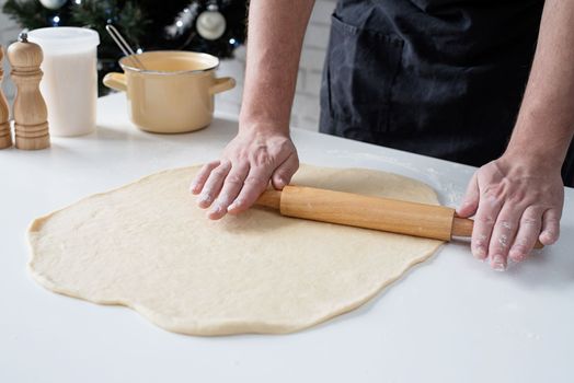 Male chef working with dough, cooking cinnamon rolls, christmas tree with lights on the background, festive cooking