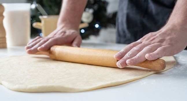 Male chef working with dough, cooking cinnamon rolls, christmas tree with lights on the background, festive cooking