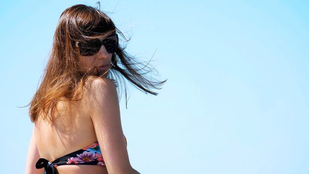 summer, sea, portrait from the back, beautiful young brunette woman wearing a bathing suit and sunglasses, enjoying the rest, her hair are fluttering in the wind. High quality photo