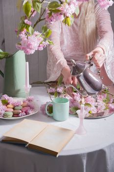 young woman pours coffee into a cup on a background of sakura flowers and colored macaroon cakes, retro style life, spring season, High quality photo 
