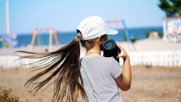 back view, teenage child, long-haired blonde girl in a blazer listening to music with bluetooth portable speaker , on the beach, dancing , on a hot summer day. High quality photo