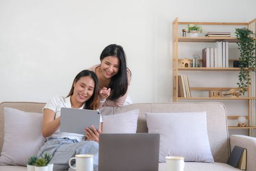 Two happy Asian women best friends in casual wear laughing while working with tablet at home in living room..
