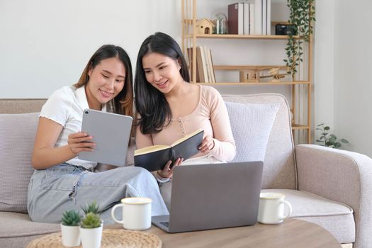 Two happy Asian women best friends in casual wear laughing while working with tablet at home in living room..