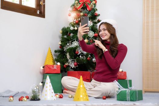 asian young woman has video chat on smartphone and shows present box to her friend with christmas tree.