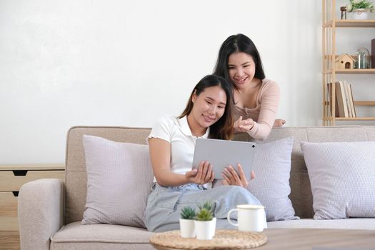 Two happy Asian women best friends in casual wear laughing while working with tablet at home in living room..